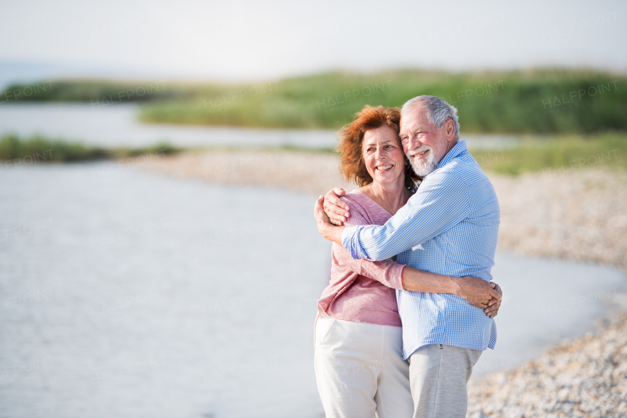 Front view of senior couple on a holiday on a walk by the lake, hugging.