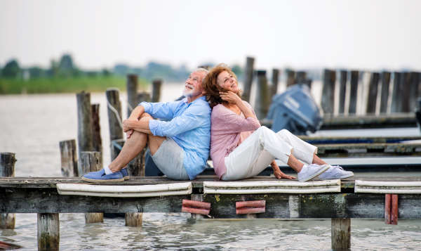 Cheerful senior couple on a holiday sitting back to back by the lake.