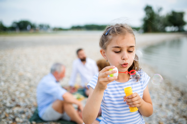 Front view of small girl with family on a holiday by the lake, making soap bubbles.