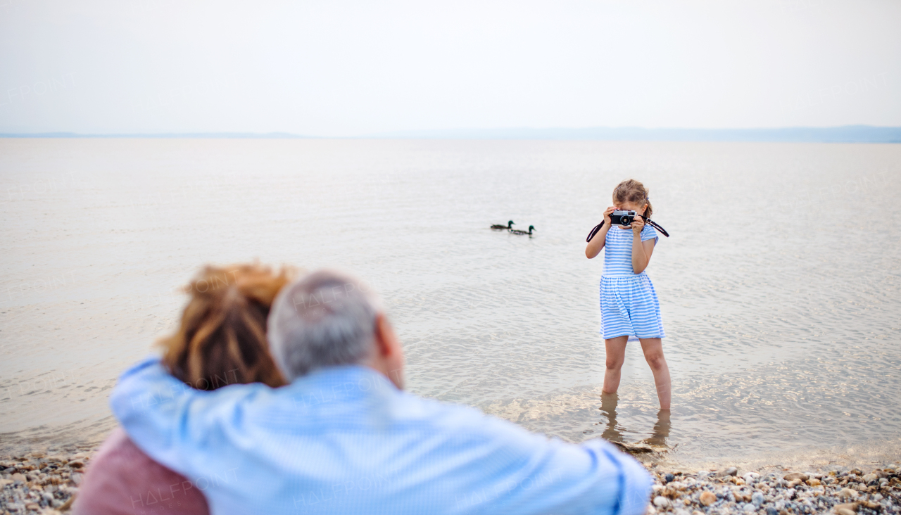 A small girl with camera and grandparents on holiday ba lake, taking photos.