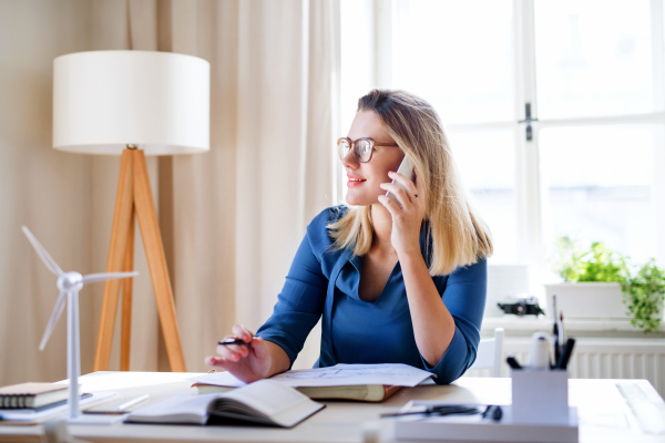 A young woman architect with smartphone sitting at the desk indoors in home office, working.