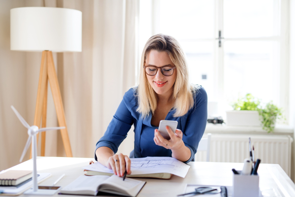 A young woman architect with smartphone sitting at the desk indoors in home office, working.