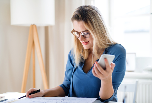 Young woman architect with smartphone sitting at the desk indoors in home office, working.