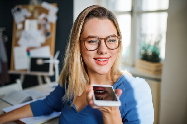 Young woman with smartphone sitting at the desk indoors in home office, making phone call when working.