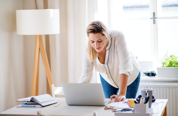 Young woman architect with laptop standing at the desk indoors in home office, working.