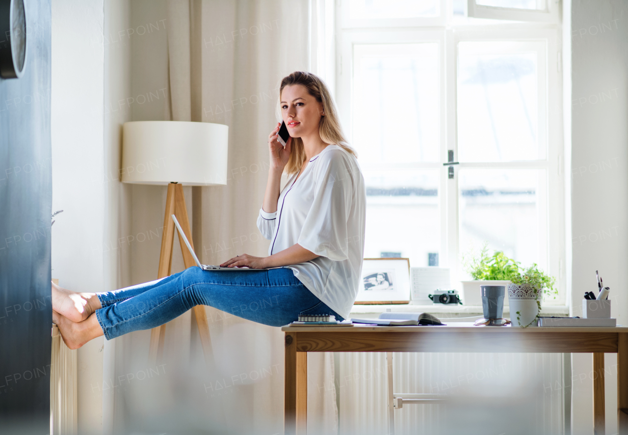 A young woman with smartphone and laptop indoors in home office, working.