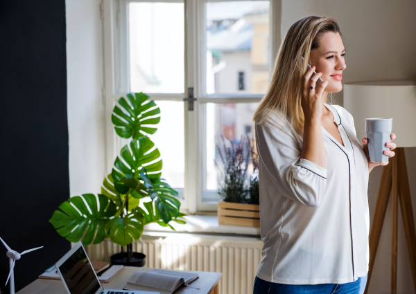 Young woman with smartphone standing indoors in home office, making phone call when working.