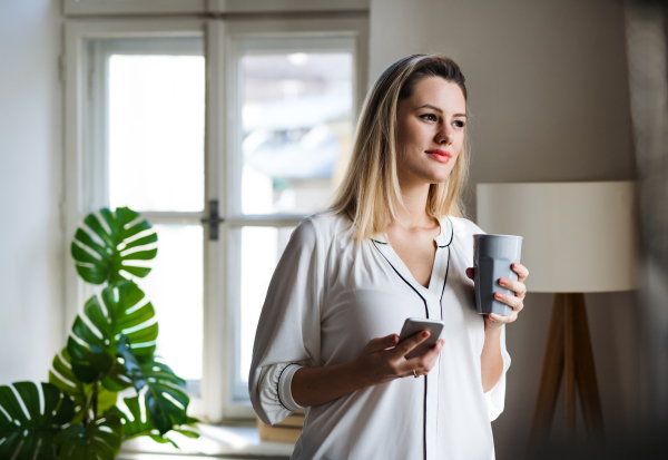 Young woman with smartphone standing indoors in home office, holding drink.