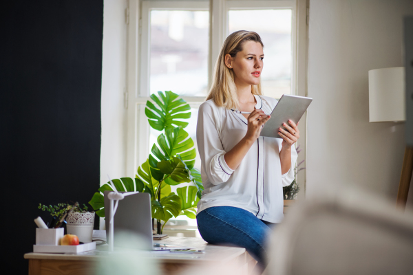 Young woman with tablet sitting on desk indoors in home office, working.