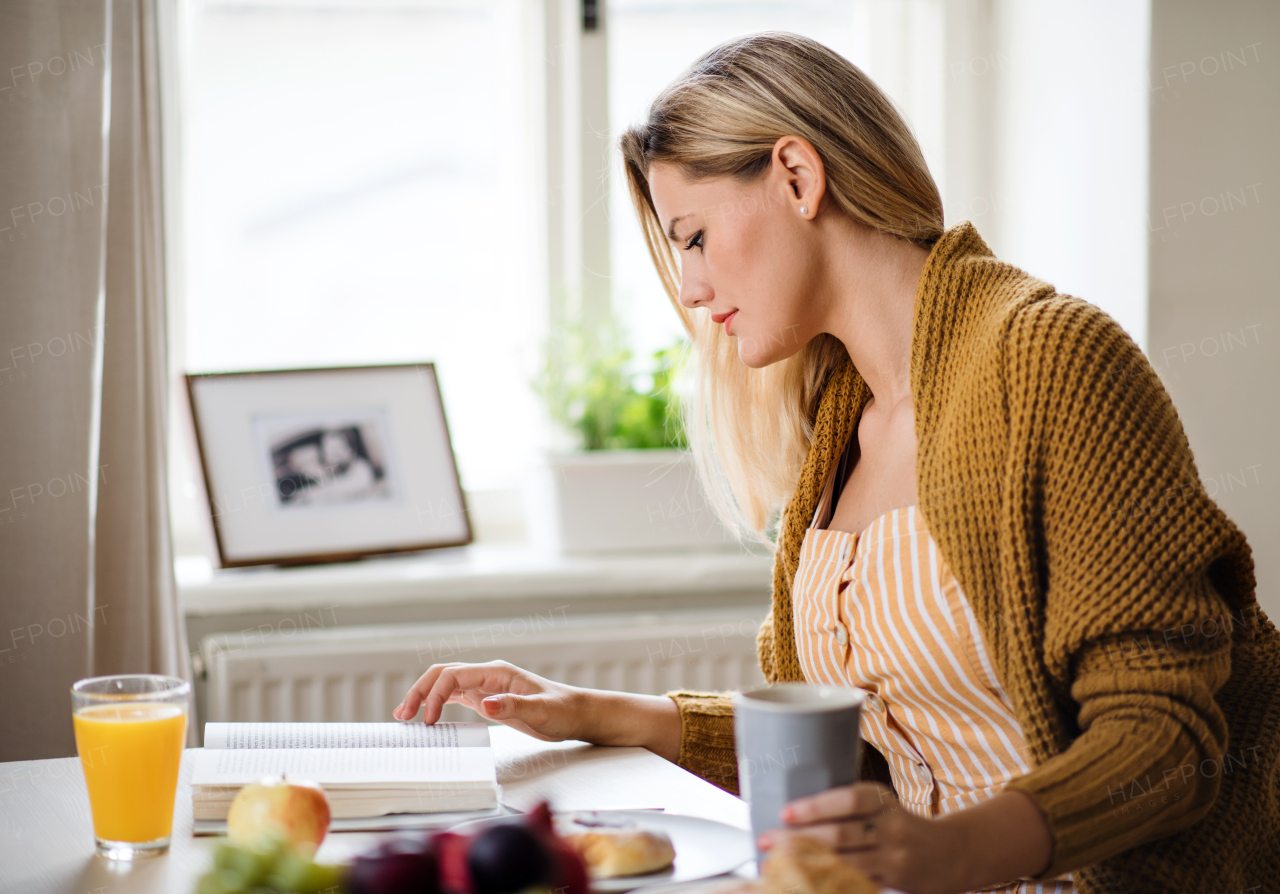 Young woman sitting at the table indoors at home, reading a book and eating.