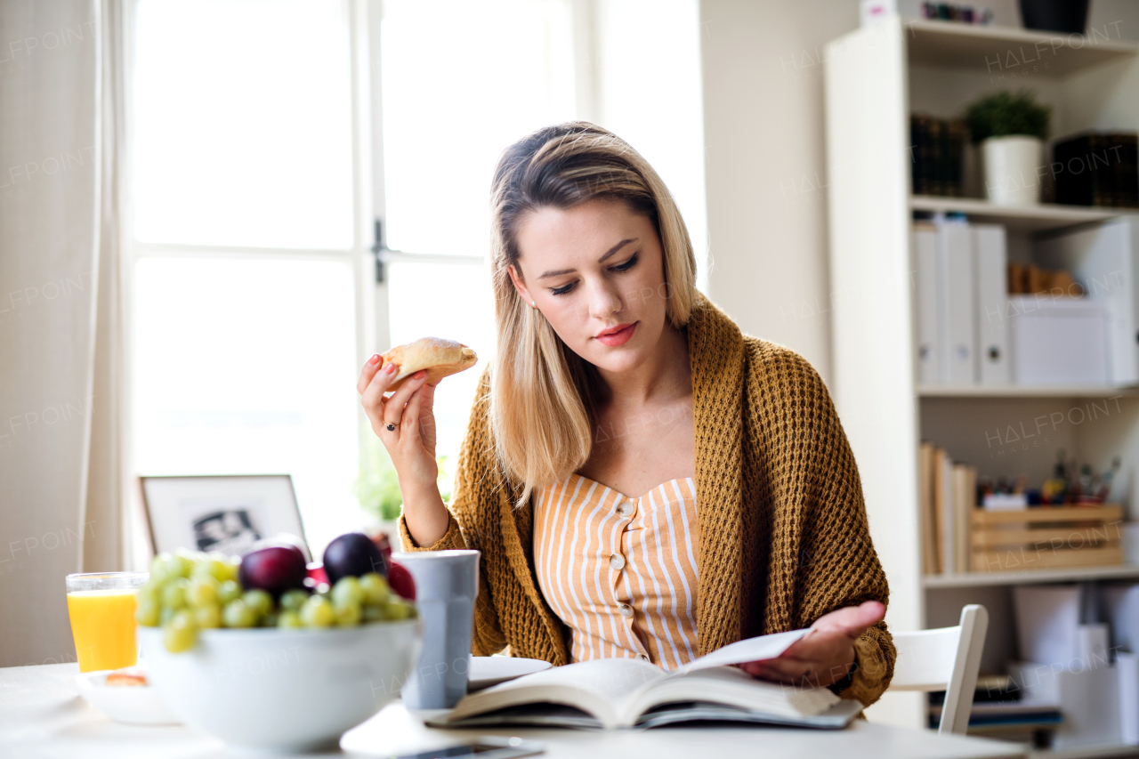 Young woman sitting at the table indoors at home, reading a book and eating.