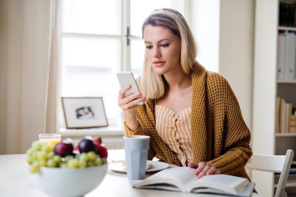 Young woman sitting at the table indoors at home, using smartphone when relaxing.