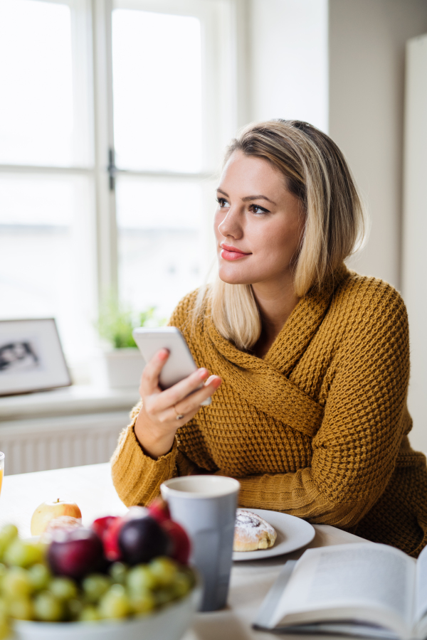 Young woman sitting at the table indoors at home, using smartphone when relaxing.