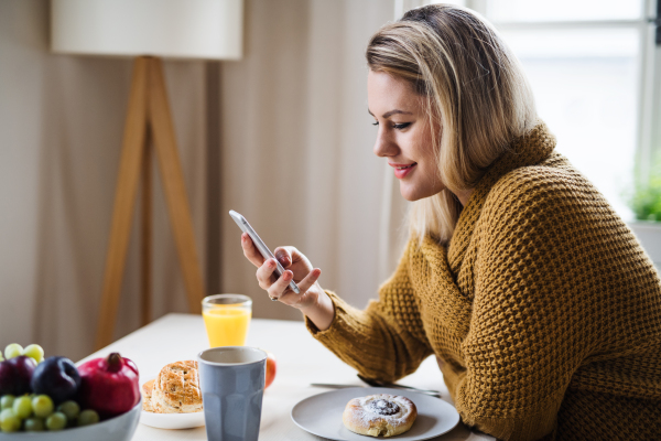 Young woman sitting at the table indoors at home, using smartphone when relaxing.