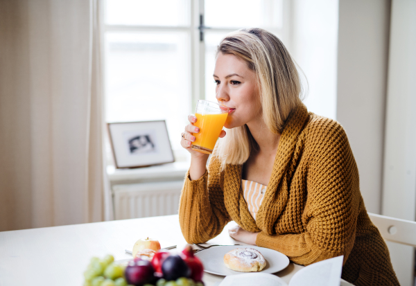 A young woman sitting at the table indoors at home, drinking juice.