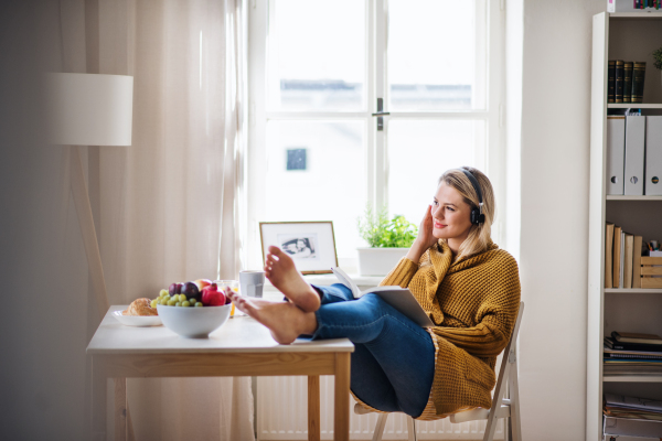 Happy young woman with headphones sitting at the table indoors at home, reading a book.