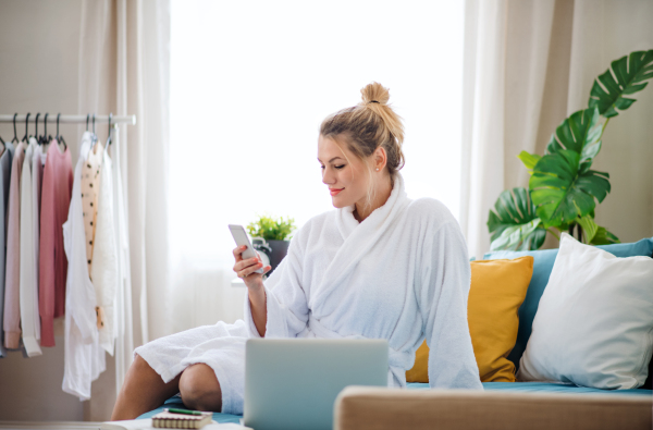 A young woman with telephone and laptop sitting on bed indoors at home in the morning.