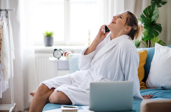 A young woman with telephone and laptop sitting on bed indoors at home in the morning.