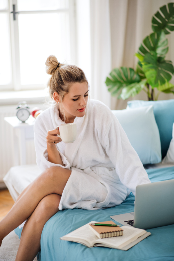 A young woman with coffee sitting on bed indoors at home in the morning, using laptop.