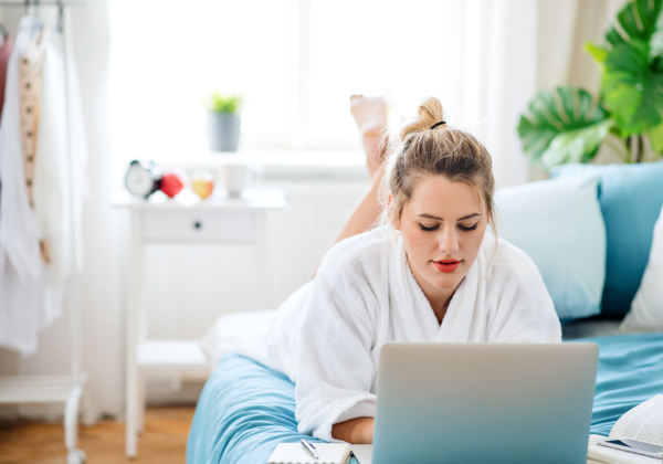 A young woman lying on bed indoors at home in the morning, using laptop.