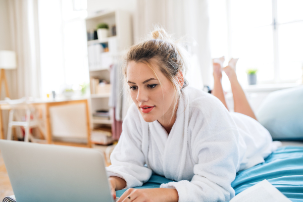 A young woman lying on bed indoors at home in the morning, using laptop.