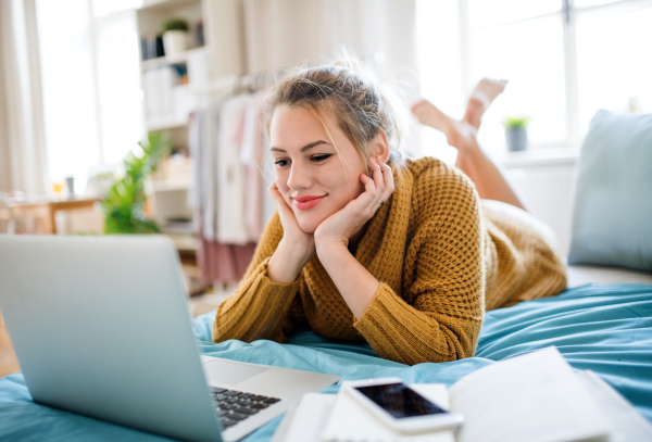 Happy young woman with laptop lying on bed indoors at home, relaxing.