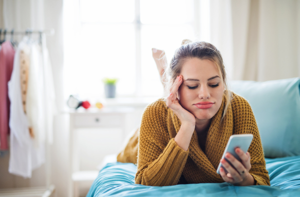 Bored young woman with smartphone lying on bed indoors at home, relaxing.