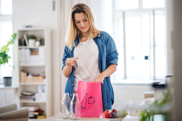 Front view of young woman putting glass bottles in bag indoors at home, recycling concept.