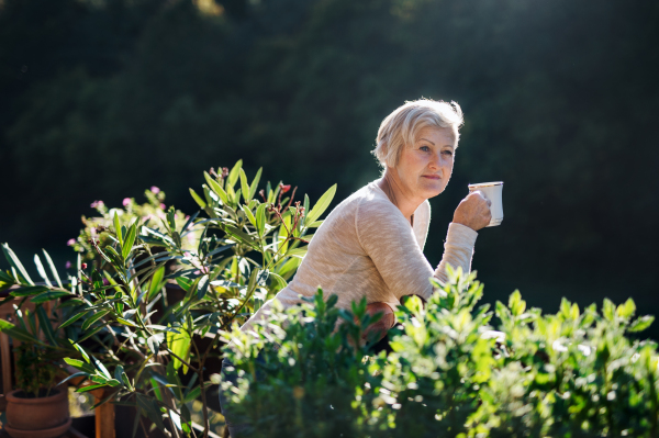 Side view of senior woman with coffee standing outdoors on terrace, resting.