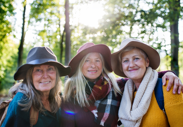 Senior women friends with hat walking outdoors in forest, looking at camera.