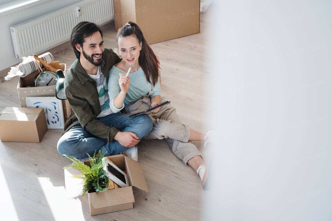 A high angle view of young couple with tablet sitting on floor and planning when moving in new flat, new home and relocation concept.