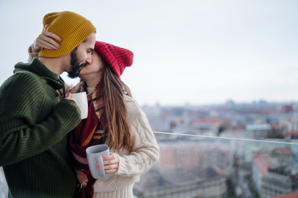 A young couple owners with tea on balcony in new flat, kissing, new home and relocation concept.