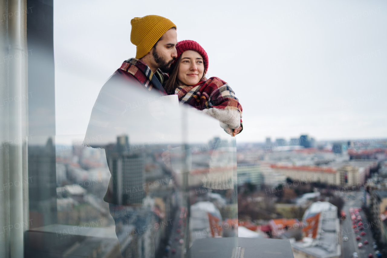 A young couple wrapped in blanket standing and hugging outdoors on balcony, shot through glass.