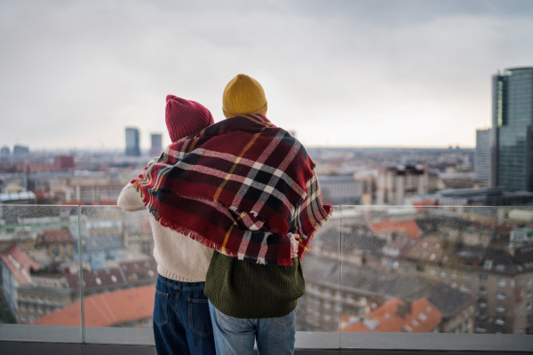 A rear view of young couple wrapped in blanket standing and hugging outdoors on balcony, looking at city view.