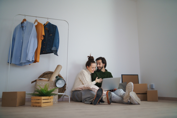 A young couple with laptop sitting on floor and planning when moving in new flat, new home and relocation concept.