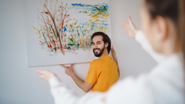 A young couple arranging new flat, hanging up painting, new home and relocation concept.