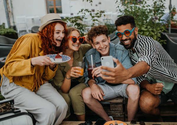 A group of happy young people with smartphone sitting in outdoors cafe on town trip, taking selfie.