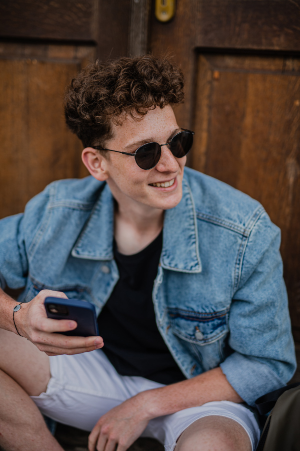 A portrait of happy young man outdoors in front of door, using smartphone.