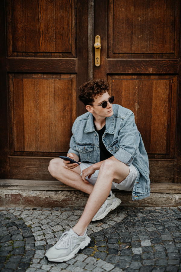 A portrait of happy young man outdoors in front of door, using smartphone.