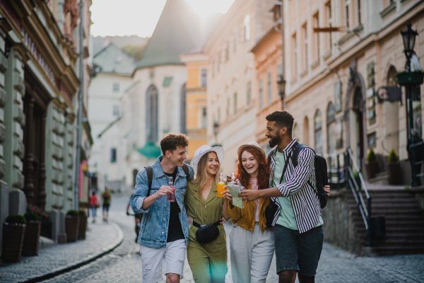 A front view of group of happy young people outdoors on street on town trip, walking with drinks.