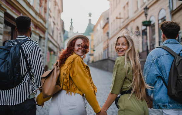 A rear view of group of happy young people outdoors on trip in town, walking and looking back.