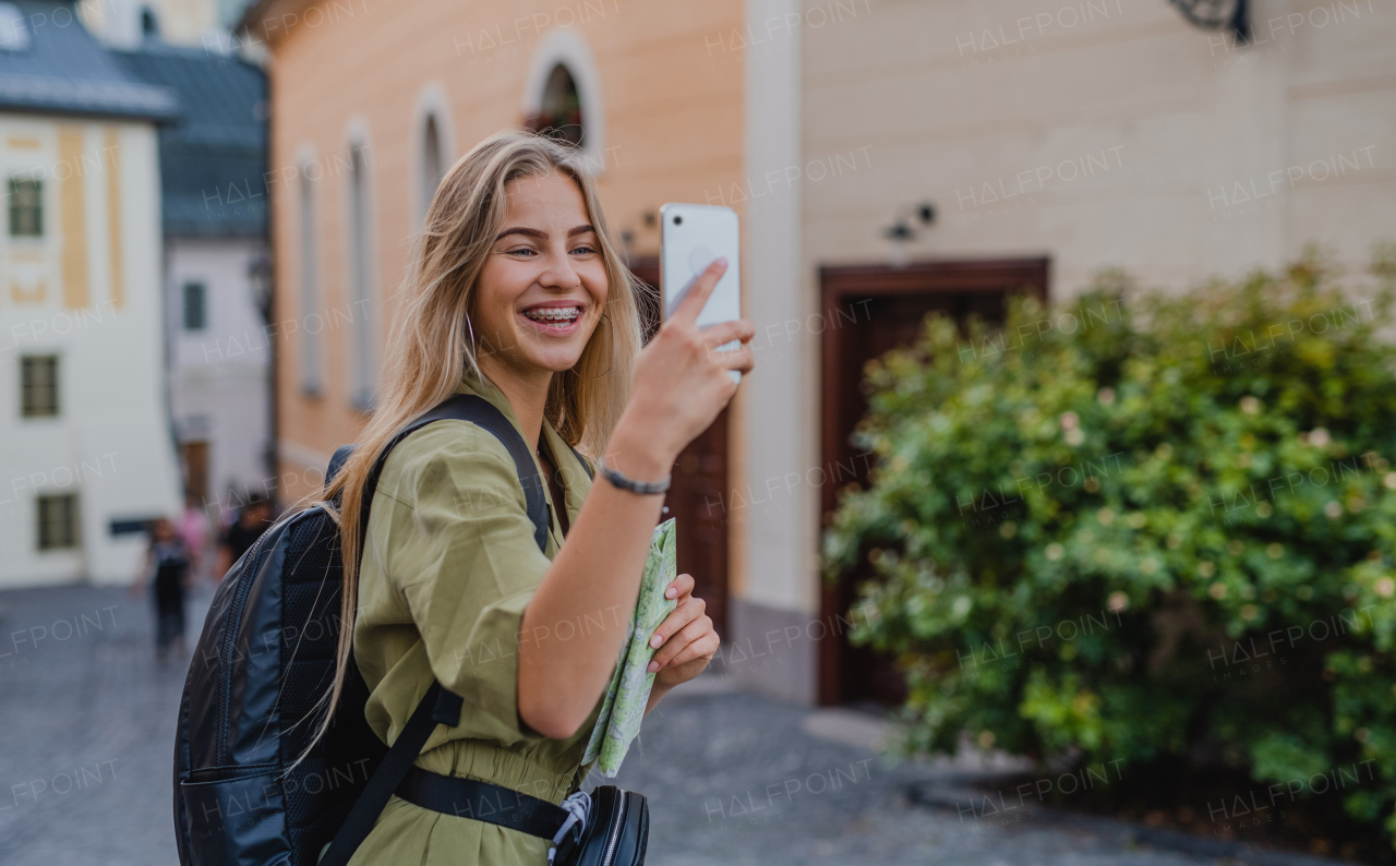 Happy young woman tourist outdoors on trip in town, taking selfie with smartphone.