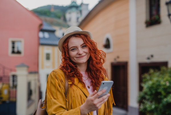 A young woman tourist with smartphone outdoors on trip in town, looking at camera.