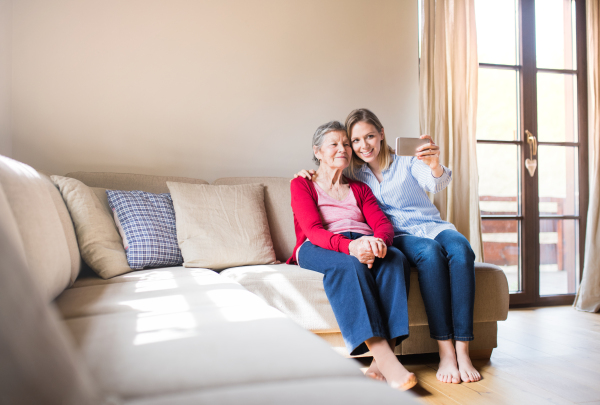 An elderly grandmother and adult granddaughter with smartphone at home, taking selfie.