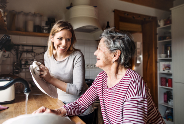 A happy elderly grandmother with an adult granddaughter at home, washing the dishes.