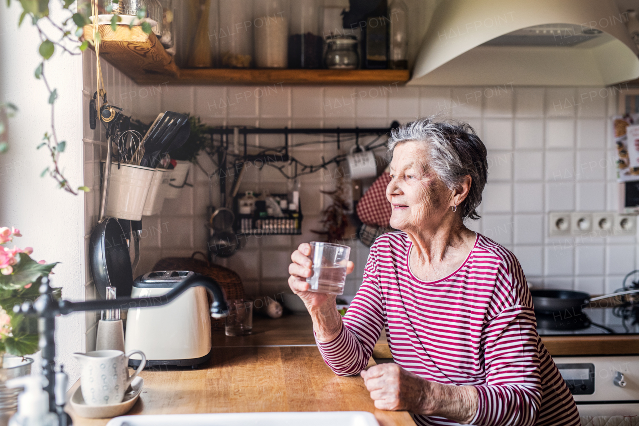 A happy elderly woman standing in the kitchen, holding a glass of water.