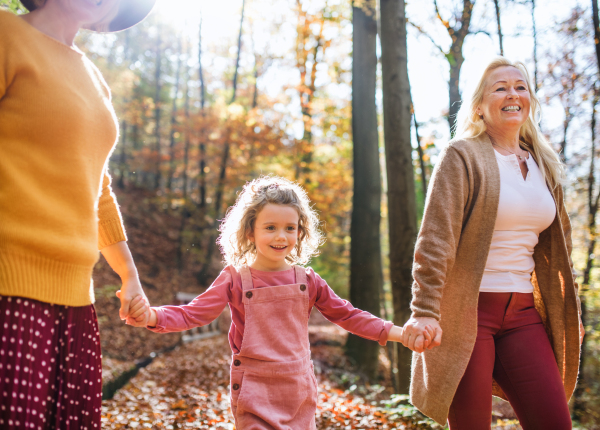 Small girl with unrecognizable mother and grandmother on a walk in autumn forest.