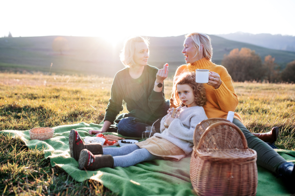 Happy small girl with mother and grandmother having picnic in nature at sunset.