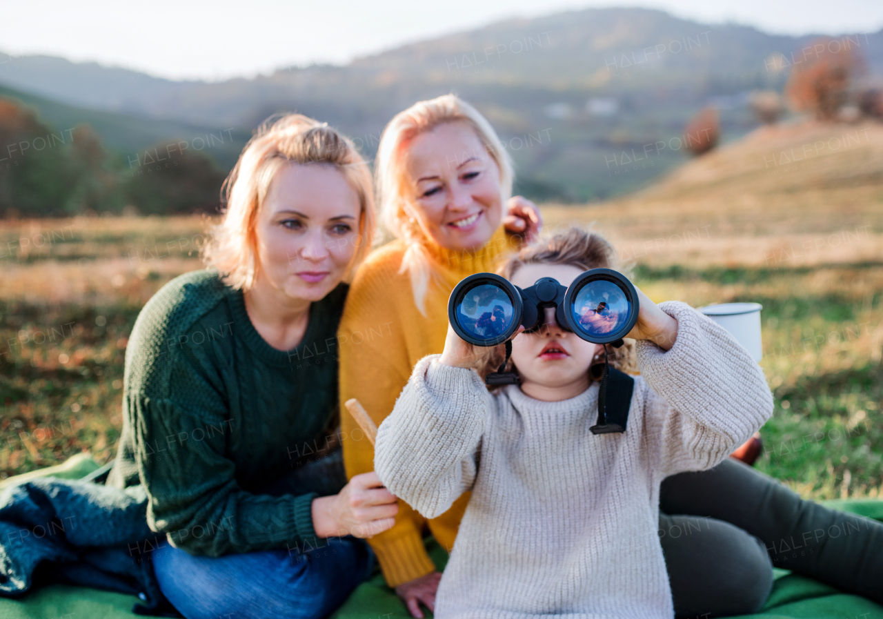 Portrait of small girl with mother and grandmother on a walk in autumn nature, using binoculars.