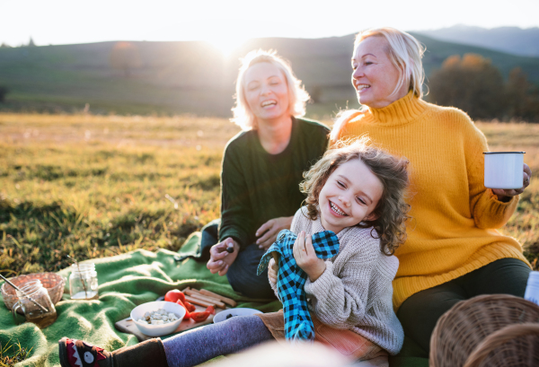 Happy small girl with mother and grandmother having picnic in nature at sunset.